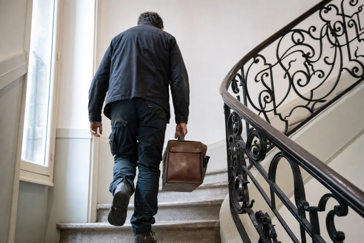 Serrurier de dos portant sa sacoche à outils dans une cage d’escalier d’immeuble lyonnais